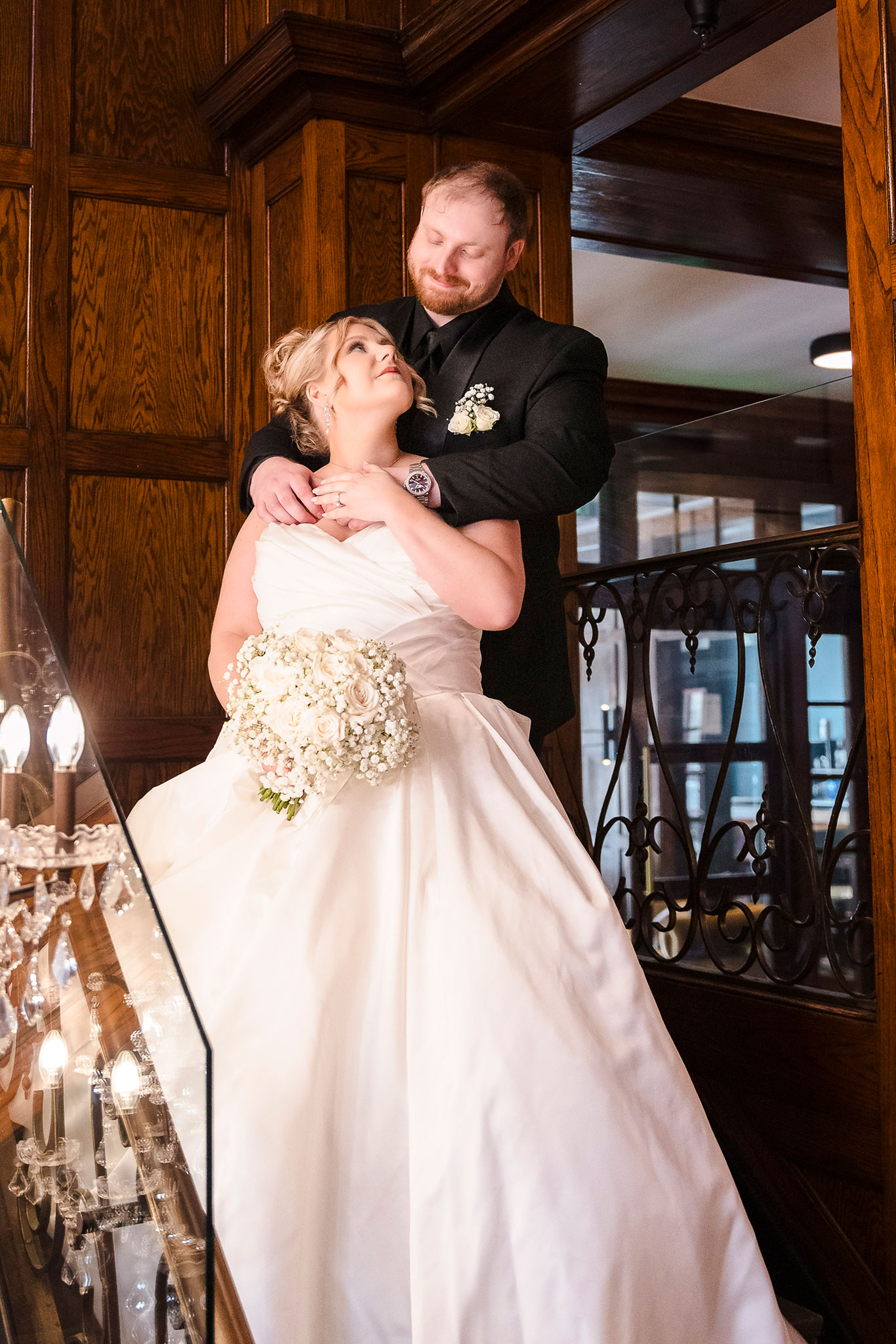Bride in a classic white gown holding a bouquet of white roses and baby’s breath, gazing lovingly up at her groom in a black suit as they embrace on a grand wooden staircase with elegant iron railings at a historic wedding venue.