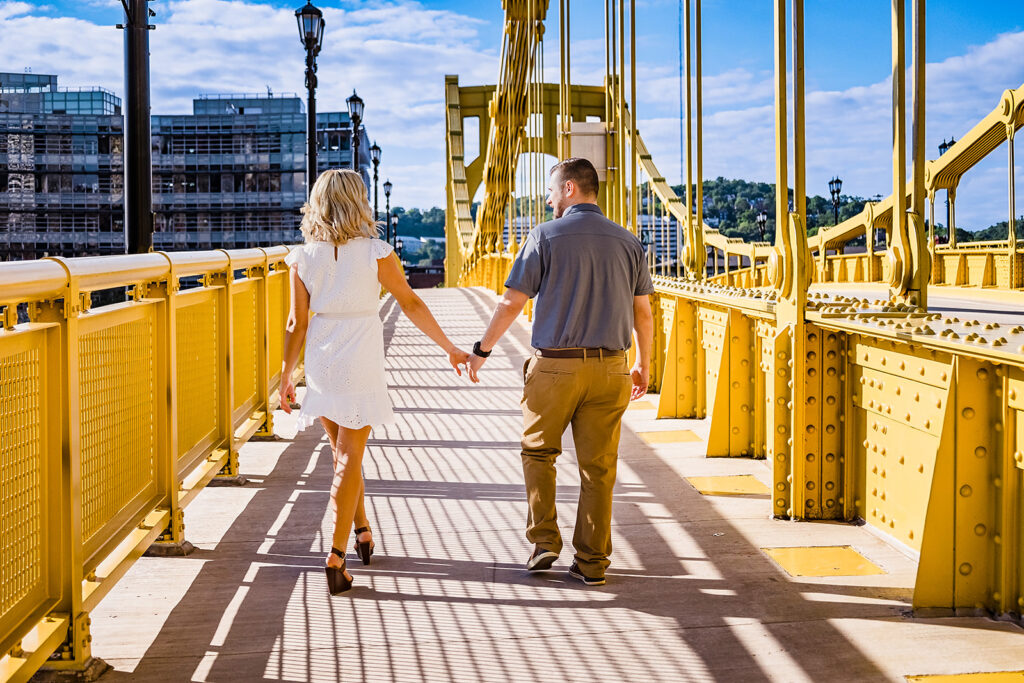 Engaged couple holding hands and walking across a bright yellow bridge in Pittsburgh on a sunny day, with the city skyline and blue sky in the background.